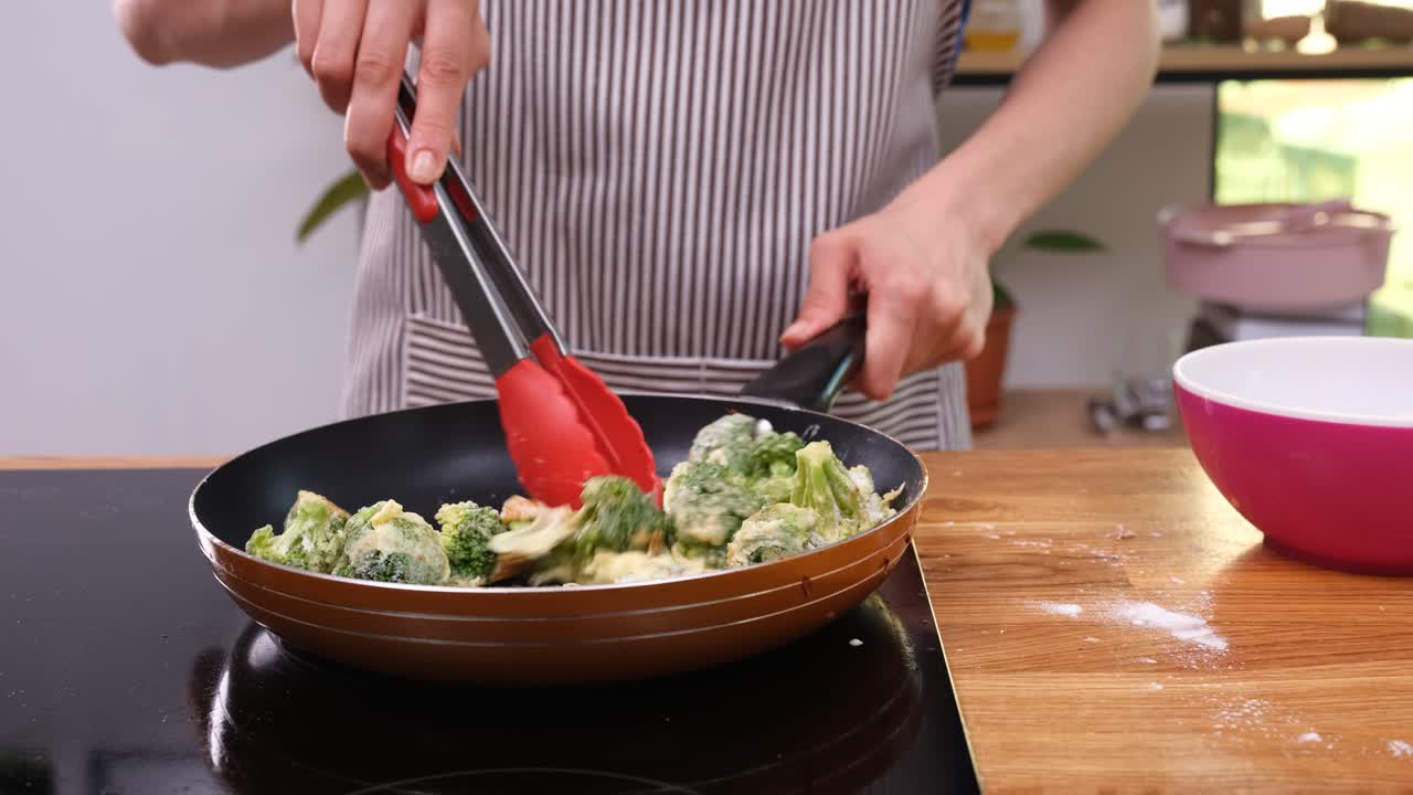 Cozinheiro vira brócolis com pinças misturando molho em frigideira quente.