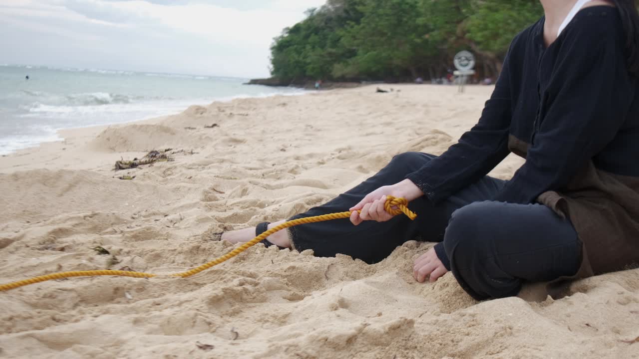 Medium shot of a young woman sitting by the sea holding a yellow rope by the beach leading to the sea, representing depression. -static shot
