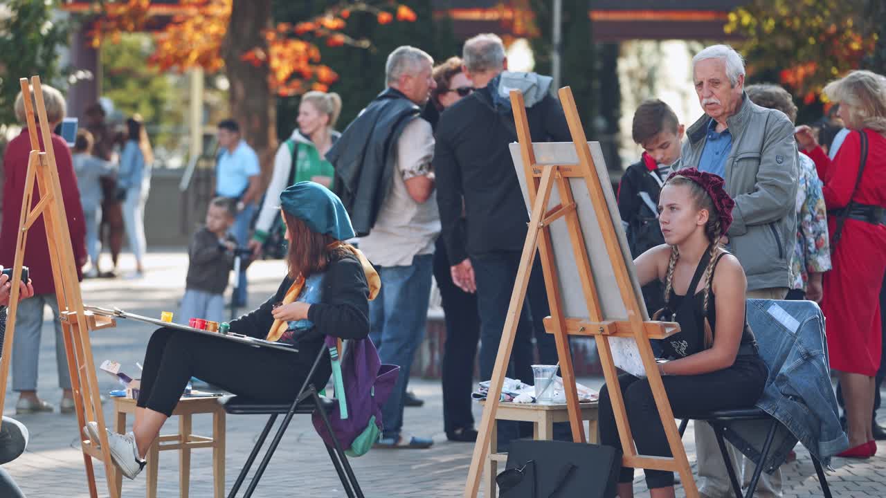 Young artists painting on easels. Artists drawing pictures on the city street with crowds of people standing nearby.