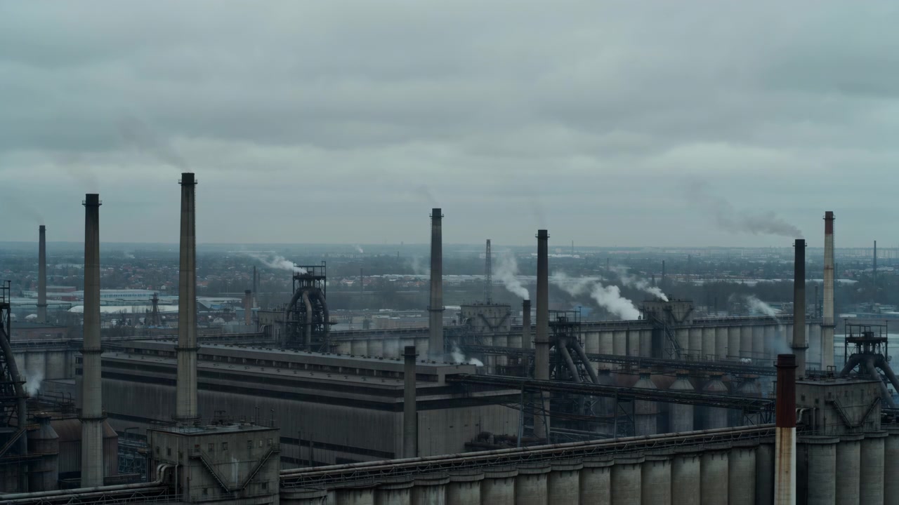 Elevated panoramic view of heavy industrial complex with tall chimneys releasing thin plumes of smoke and steam that rise and drift in light wind beneath layered gray clouds and muted overcast light