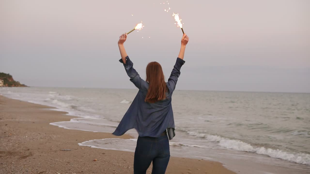 vista posterior de una joven mujer atractiva corriendo por el mar durante la puesta del sol y sosteniendo velas brillantes en ambas manos