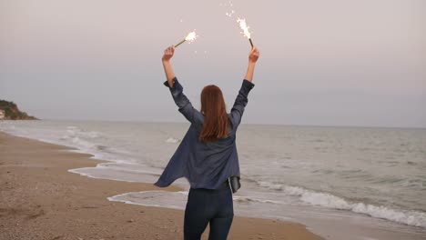 Back-view-of-young-attractive-woman-running-by-the-sea-during-sunset-and-holding-burning-sparkling-candles-in-both-hands