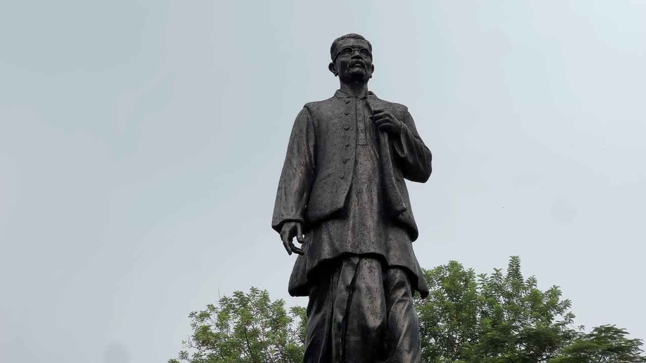 A tilt up shot of black statue of Hemant Kumar Basu surrounded by trees all around under the blue sky.