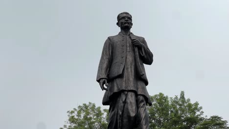 a tilt up shot of black statue of hemant kumar basu surrounded by trees all around under the blue sky.