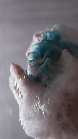 woman crumples foamy loofah in shower cabin closeup. lady shows mesh body washing sponge against falling water flow. skincare and massage tool