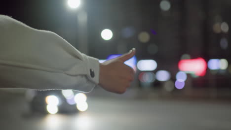 female hand in white long sleeve extended with thumb up gesture to stop taxi under bright city streetlights at night while car drives past in blurred colorful background with glowing bokeh