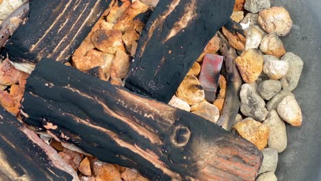 Charred-logs-over-pebbles,-beach-campfire,-close-up-slow-motion