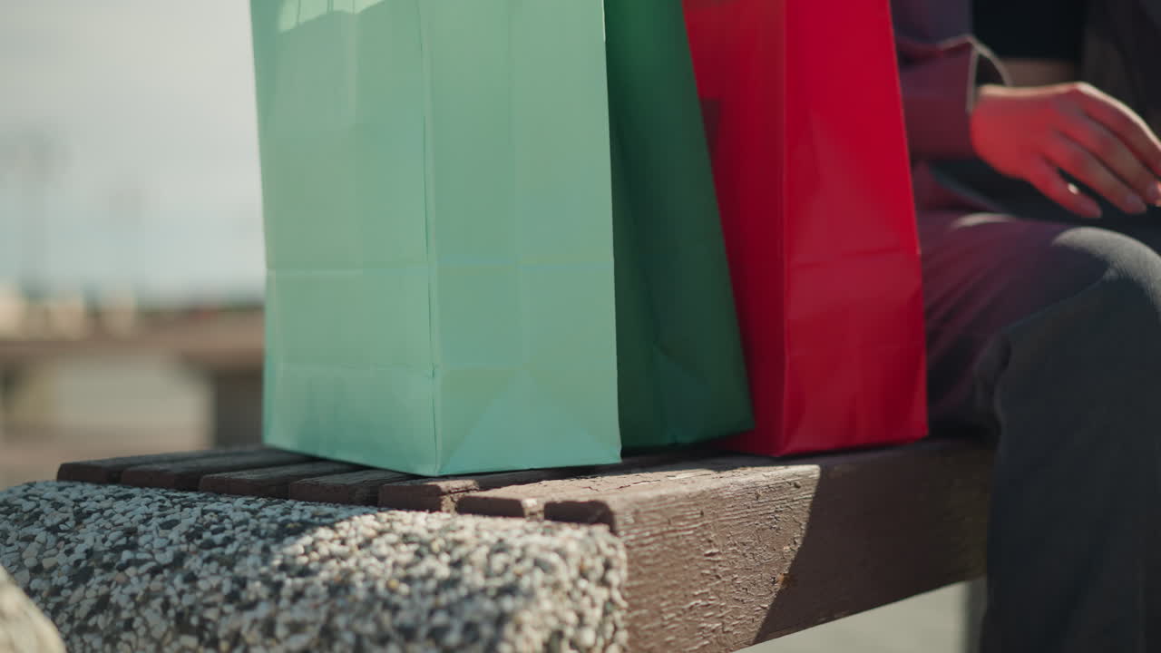 앉아있는 동안 벤치에 다채로운 쇼핑 가방을 떨어뜨리는 개인의 클로즈업 다리 뷰, 다리에 부분적으로 손을 고 있습니다.