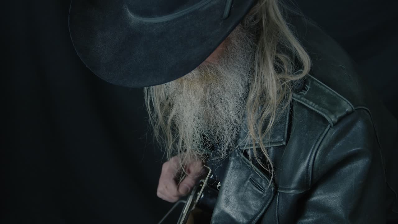 Close up bearded cowboy in black Stetson plays electric guitar in dark