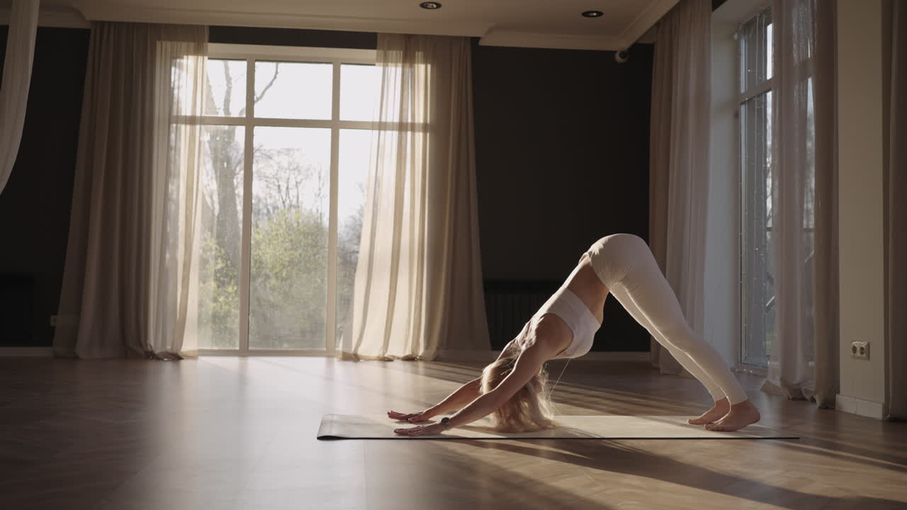 A young woman in a white suit in a gym trains yoga in the sun in slow motion