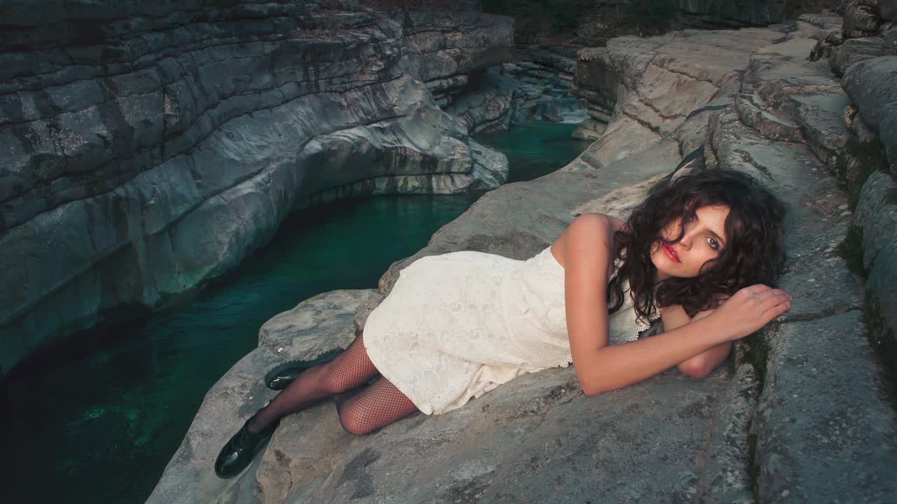 Cinemagraph - seamless video loop of a young brunette model girl in a white dress sitting on the edge of a small canyon by a blue mountain river. The water is rushing through naturally formed rocks.