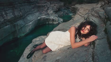 cinemagraph - seamless video loop of a young brunette model girl in a white dress sitting on the edge of a small canyon by a blue mountain river. the water is rushing through naturally formed rocks.