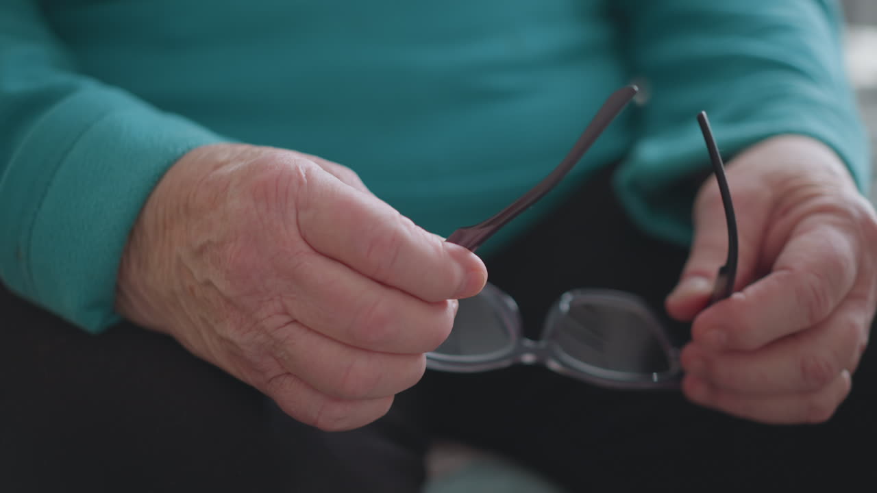Elderly woman in green teal sweater adjusting eyeglasses while sitting. She focuses carefully as she holds glasses, possibly preparing to wear them, in a cozy home setting