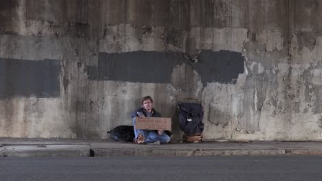 Wide-Shot-of-a-Homeless-Man-Sitting-on-the-Sidewalk-Holding-a-God-Bless-Cardboard-Sign-Begging-for-Money