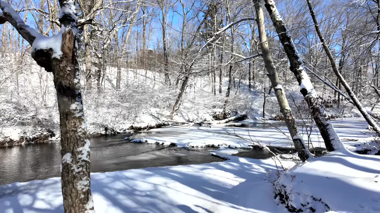 A peaceful winter scene featuring a flowing creek surrounded by snow-covered trees. The icy water reflects the sunlight, adding a serene and magical touch to the snowy forest landscape