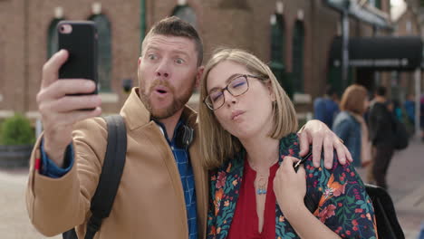 portrait-of-happy-couple-embrace-posing-making-faces-taking-selfie-photo-using-smartphone-in-city