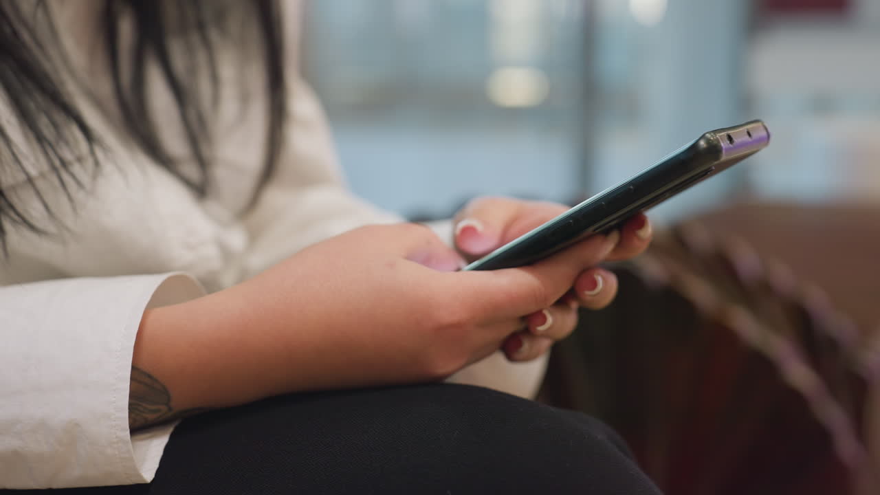 Close up side view of young woman in white shirt using smartphone with both hands while seated indoors, black pants visible, phone screen reflecting soft light