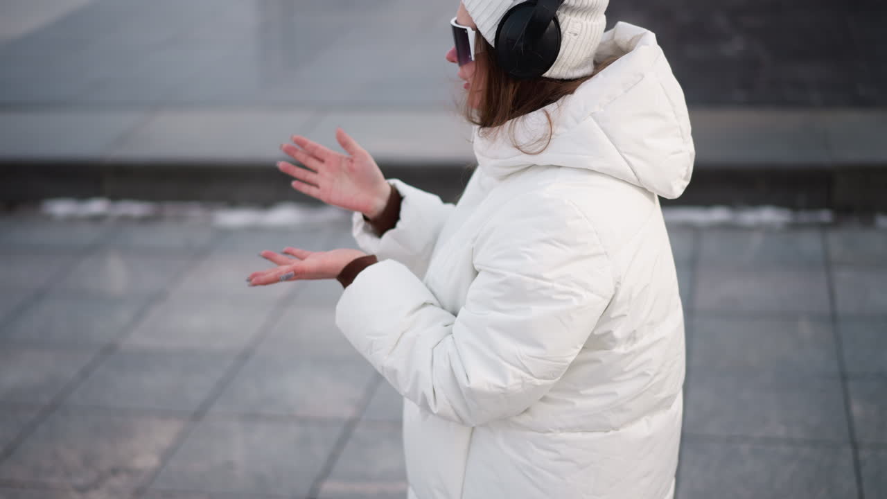 Elegant young lady wearing headset moving hands as morning routine outdoors on tiled pavement in winter coat under pale sky backdrop with focused expression, casual energy and rhythmic motion