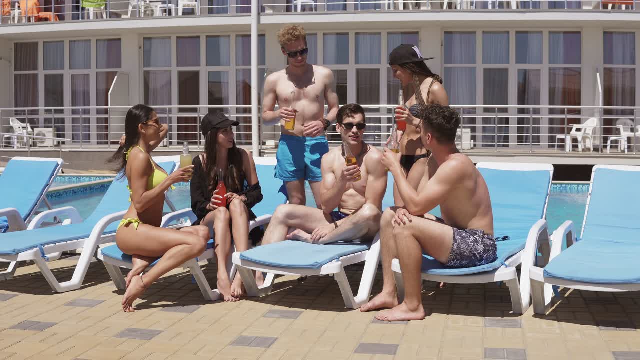 grupo de hermosos amigos jóvenes bebiendo cócteles y divirtiéndose sentados junto a la piscina. rodado en 4k