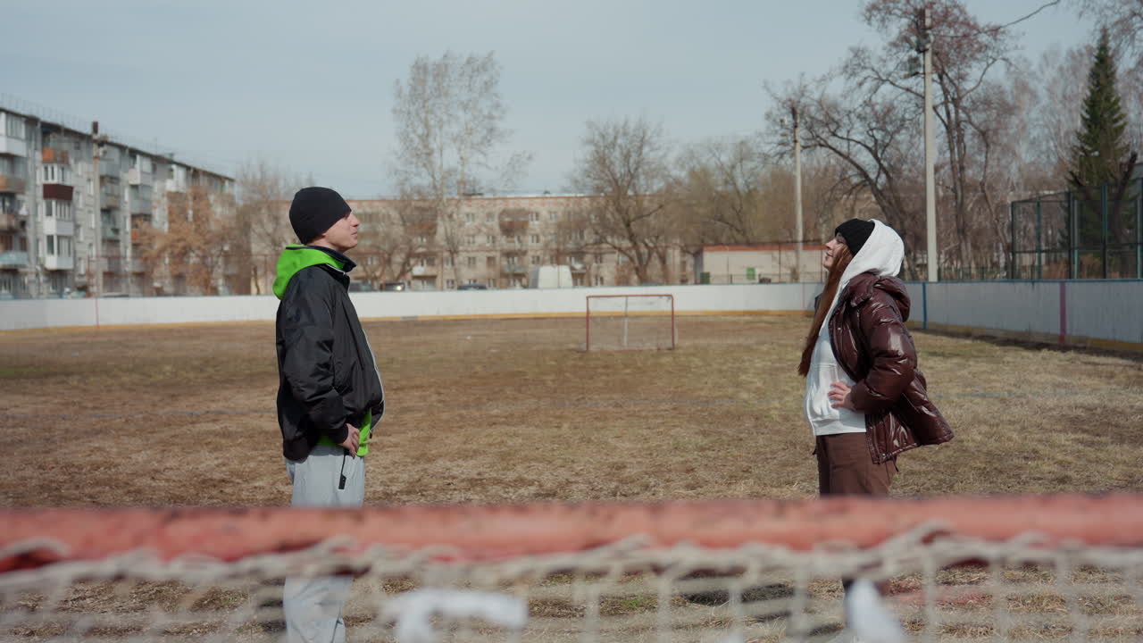 텅 빈 경기장에서 마주 보고 서 있는 백인 십 대들, 전경에는 골대, 뒤로는 아파트 스카이라인, 진지한 대화와 몸짓, 차가운 조명과 잠재적 화해를 담은 솔직한 대립