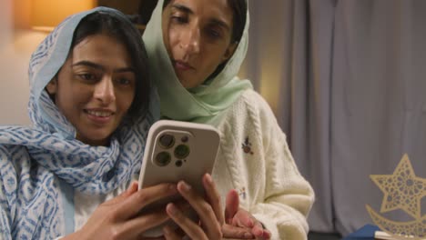 madre e hija riéndose del teléfono móvil sentadas alrededor de una mesa familiar musulmana en casa comiendo comida iftar rompiendo el ayuno diario durante el ramadán