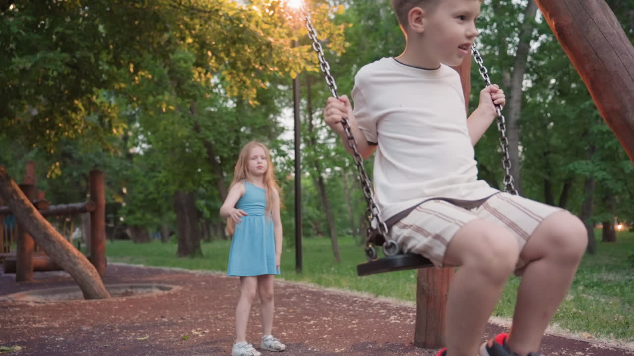 teenage boy smiles riding wooden chain swing while energetic sister pushes from behind in green park at golden dusk capturing playful sibling moment under soft warm light among trees and open air
