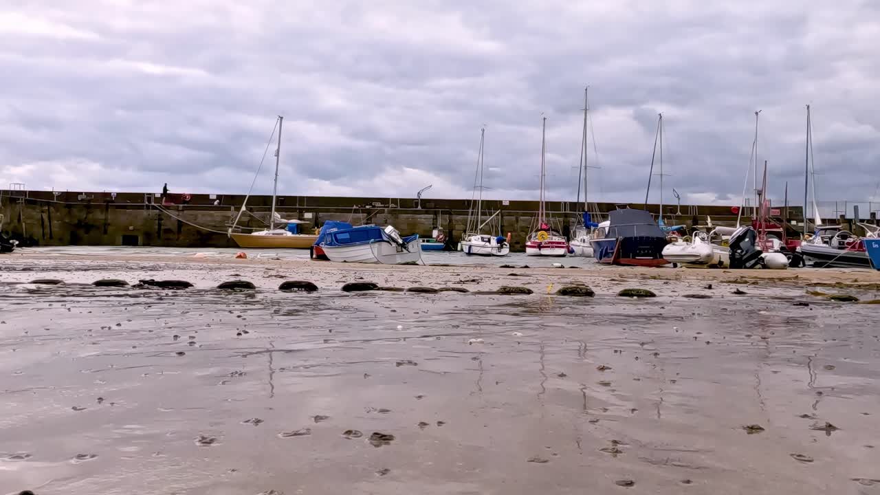 Small boats anchored on a sandy beach with cloudy skies and distant grassy hills.