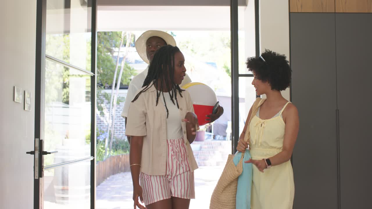 African American friends entering open glass doors, hugging, holding beach ball, carrying woven bag