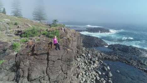 aerial drone flying forward pass woman sitting on rock cliff to kiama rock pool on a sunny misty morning with waves breaking on rocks