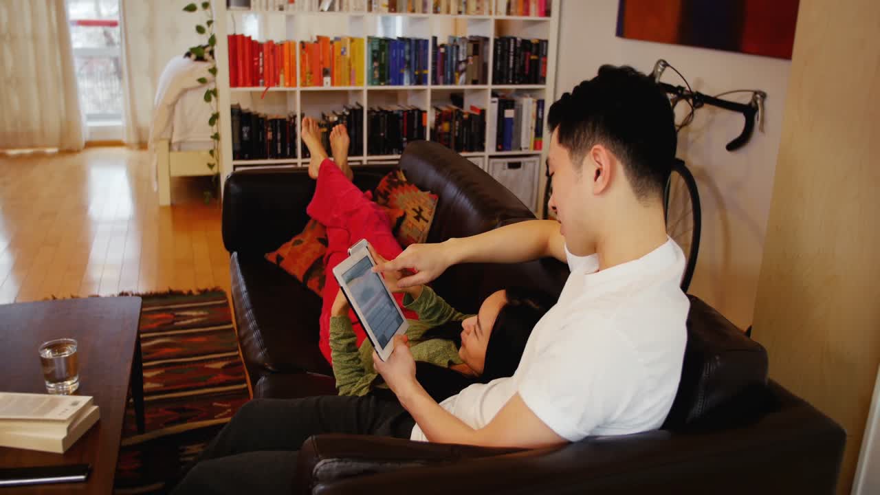 부부가 거실에서 디지털 태블릿과 휴대폰을 사용하여 소파에서 휴식을 취하고 있습니다.