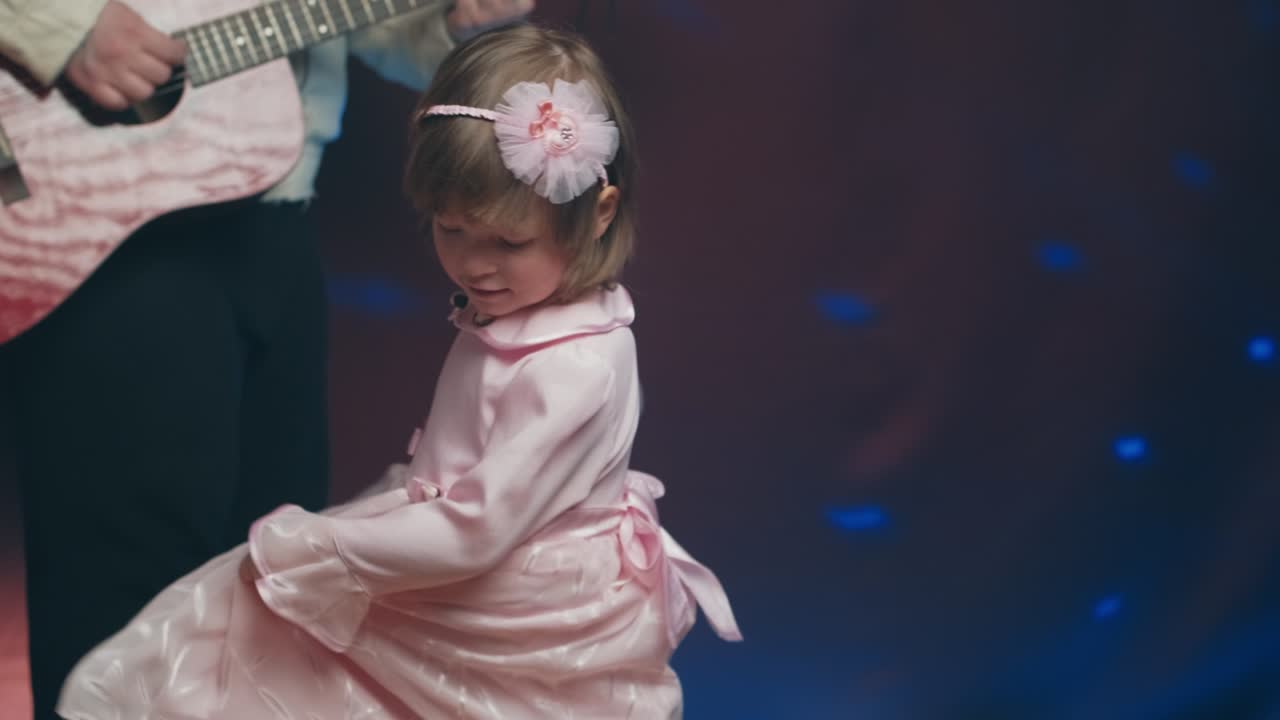 una niña con un vestido vintage baila en el escenario, su padre toca la guitarra acústica