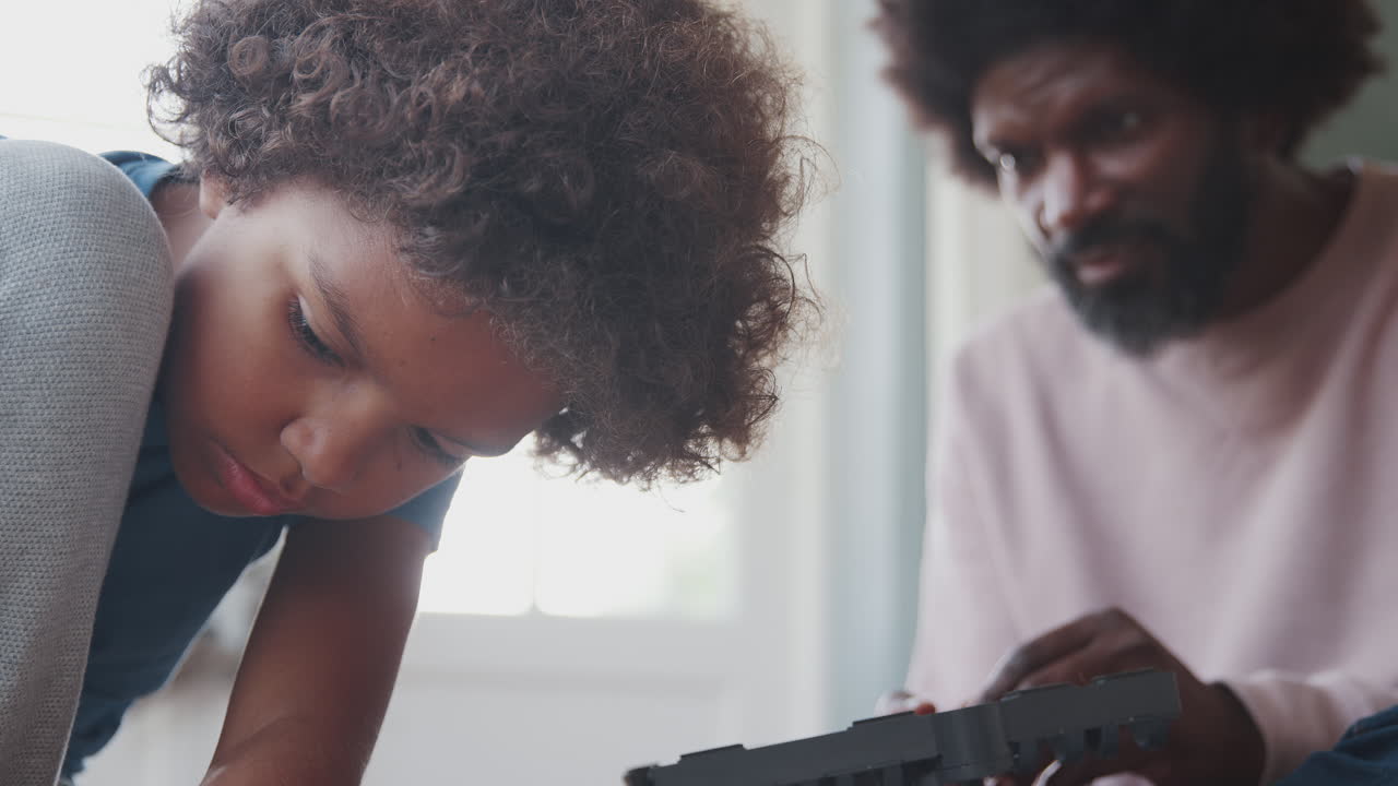 바닥에 앉아서 아버지의 지침으로 장난감을 만드는 데 집중하는 사춘기 전 소년의 클로즈업, 낮은 각도, 전면에 초점을 맞추십시오