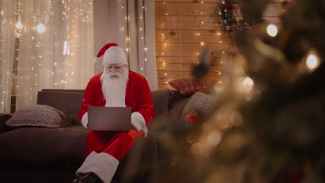 Der-Weihnachtsmann-Arbeitet-Von-Zu-Hause-Aus,-Sitzt-Auf-Der-Couch-Mit-Einem-Laptop-In-Der-Nähe-Des-Weihnachtsbaums-Und-Arbeitet-Mit-Einem-Laptop