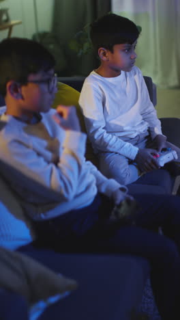 Vertical-Video-Of-Two-Young-Boys-Sitting-On-Sofa-At-Home-Playing-With-Computer-Games-Console-On-TV-Holding-Controllers-Late-At-Night