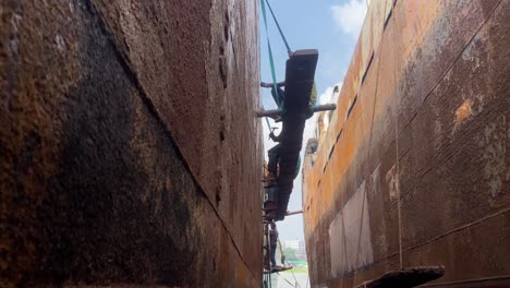 Looking-Up-At-Dock-Workers-Sitting-On-Plank-In-Between-Two-Ships-Hammering-Side