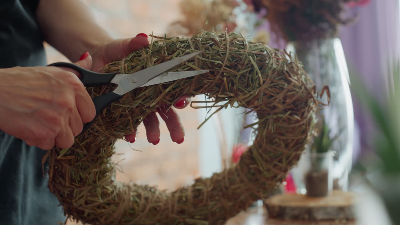 Closeup of woman trimming straw wreath with black scissors, showing detailed handcraft process, rustic decoration creation, precision cutting of natural material, artisan making floral base