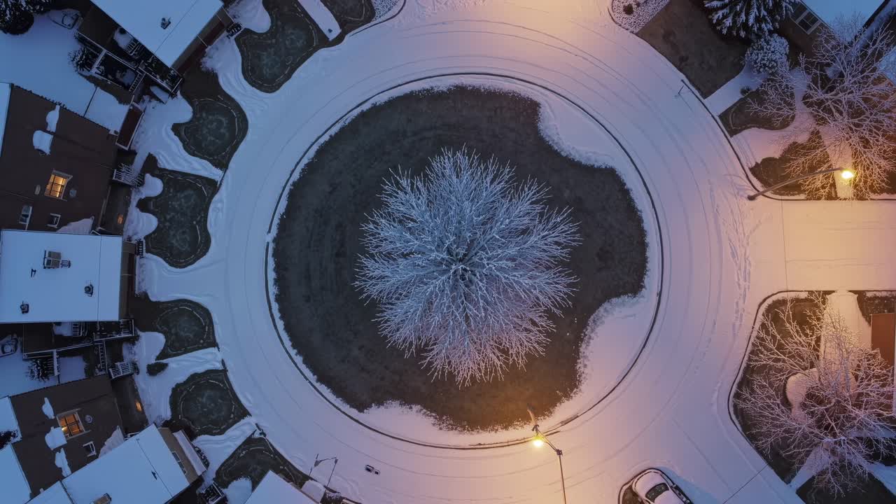Luftperspektivet fanger en sirkelformet park dekket av snø, med et sentralt tre og omkringliggende hus, og skaper en rolig vinterscene med varmt gatelys.