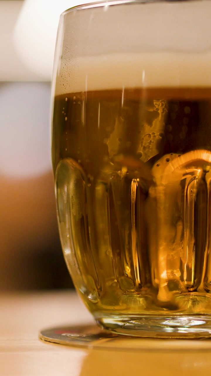 Caneca de vidro de cerveja lager dourada com colarinho de espuma, iluminação interna quente, câmera parada.