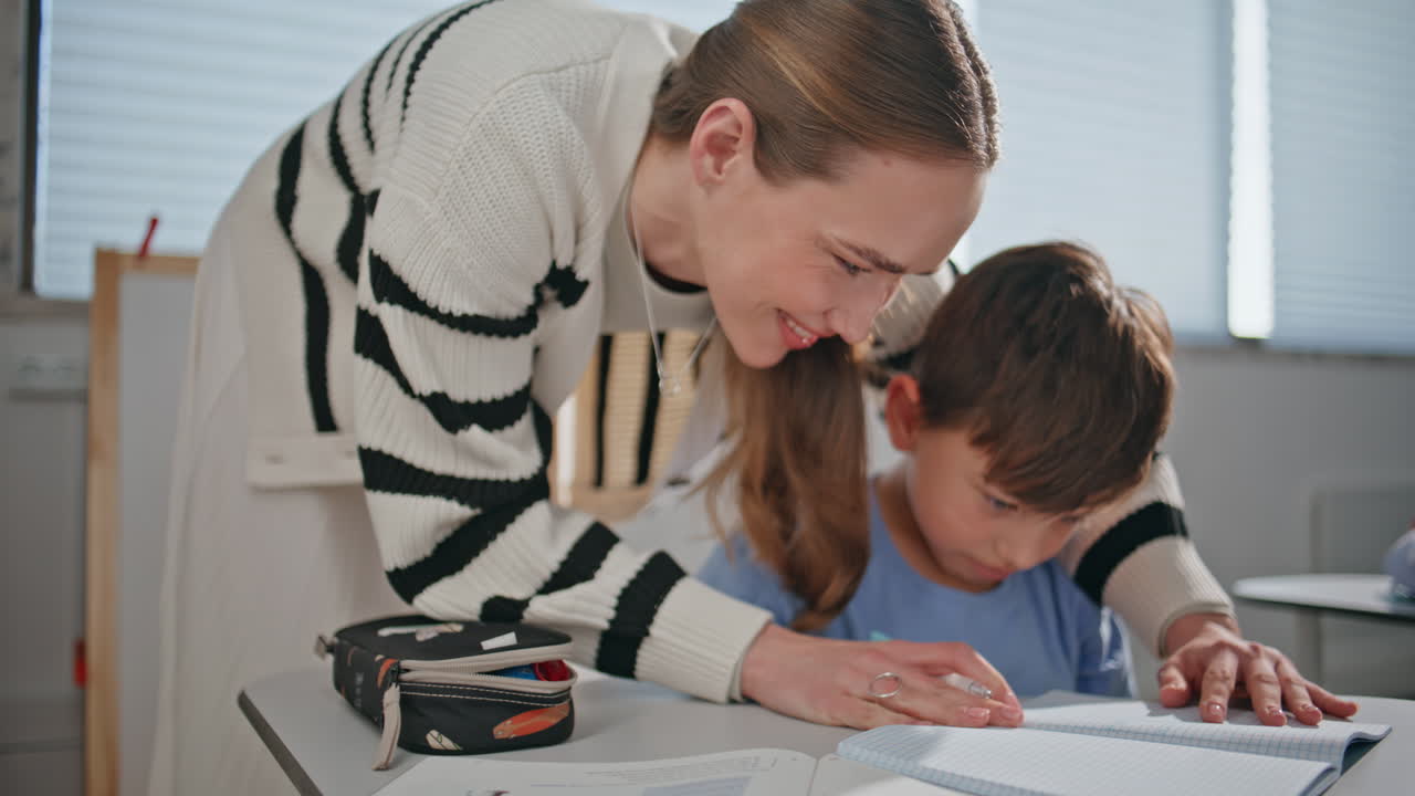 Schoolteacher child writing copybook at school. Lady helping small pupil kid