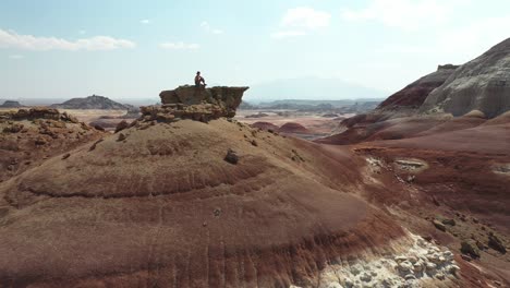 Tilt-Up-Aerial-View,-Lonely-Man-Sitting-on-Top-of-Sandstone-Hill-in-Utah-Desert-USA-With-Amazing-Lookout-on-Desert-Landscape