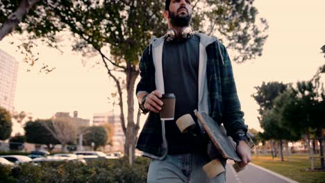 young hipster skateboarder drinking coffee and walking in urban park