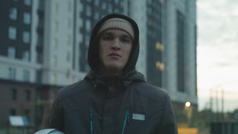 bored young man in hoodie and beanie juggling football during urban training session, showing casual expression against backdrop of tall residential buildings and cloudy sky