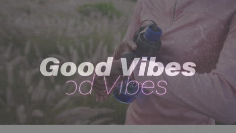 animation of the words good vibes in white over woman exercising in countryside checking watch