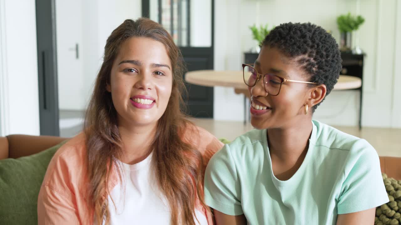 Duas mulheres de diferentes etnias trocando olhares em um sofá verde em uma sala iluminada, rindo e se apoiando, trocando carinho
