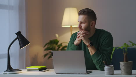 pensive man is working with laptop at home at evening writing message in online chat and working on book young guy with notebook in room