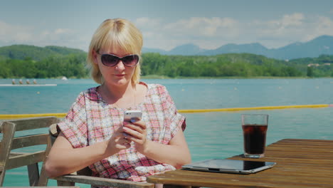 business woman uses a smartphone on vacation sitting at a table in a cafe near the lake on a backgro