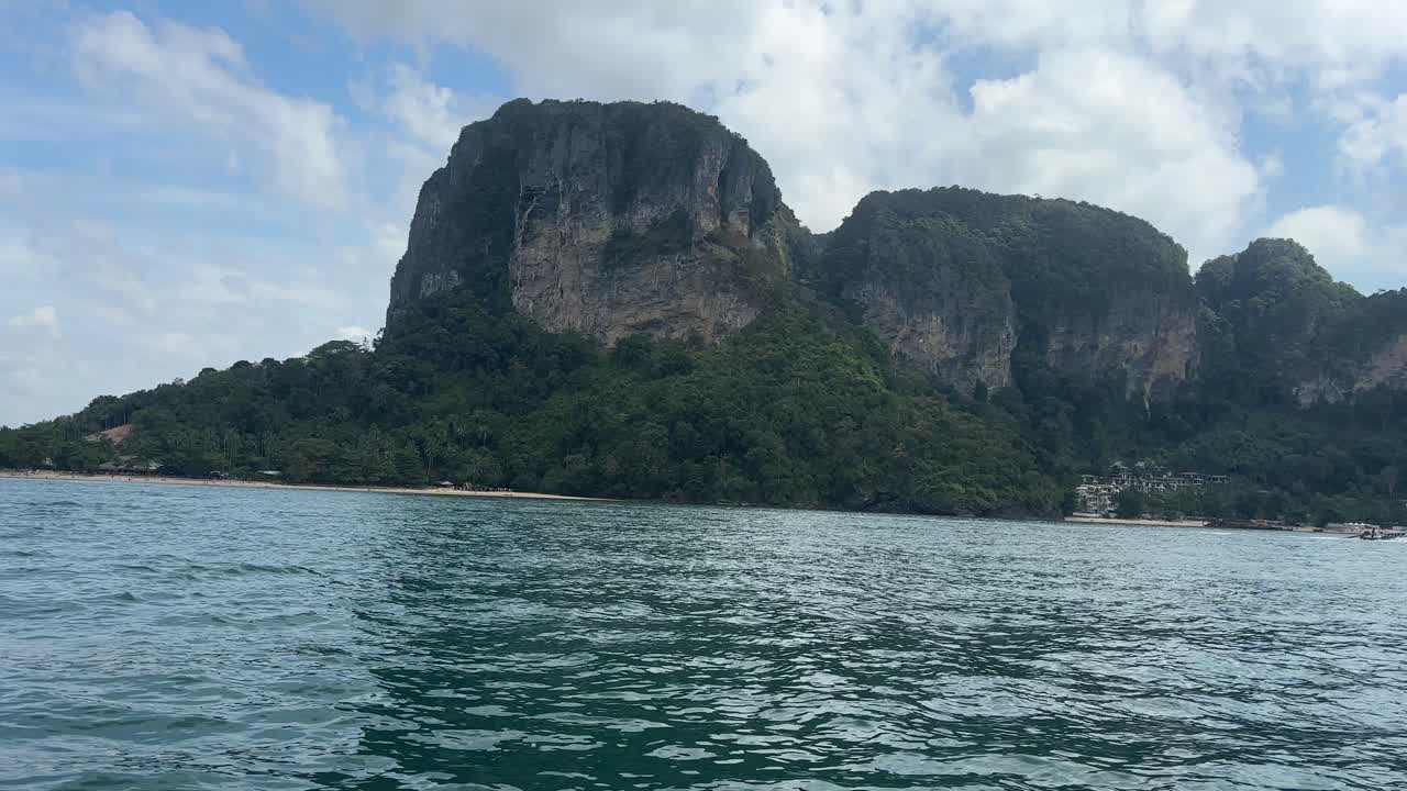 Utsikt från havet vid infarten till Railay Beach i Krabi, Thailand, som visar lugnt havsvatten i förgrunden och skogsklädda kalkstensklippor som reser sig längs den tropiska kustlinjen.