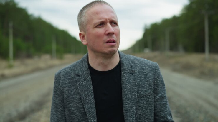 A close view of an aged white man looking focus, wearing a grey suit and a black polo, walking on a railway with stones, the background includes a blur of trees and electric poles on either side