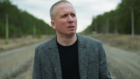 a close view of an aged white man looking focus, wearing a grey suit and a black polo, walking on a railway with stones, the background includes a blur of trees and electric poles on either side