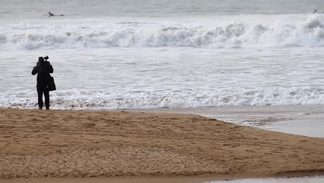 Un-Hombre-Está-De-Vacaciones-Y-Captura-La-Belleza-De-La-Playa-Antes-Del-Amanecer-Con-Su-Cámara,-Tomando-Fotos-Del-Océano-Con-Olas-Rompiendo-En-El-Fondo